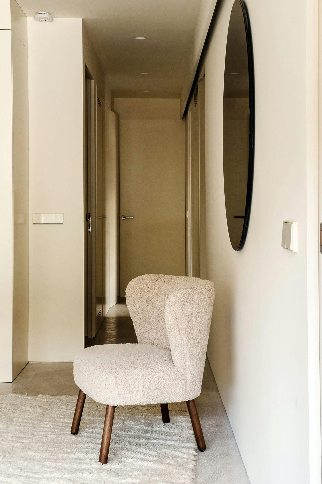 A white chair is sitting in a hallway, with a mirror above it, in a clean and empty room.