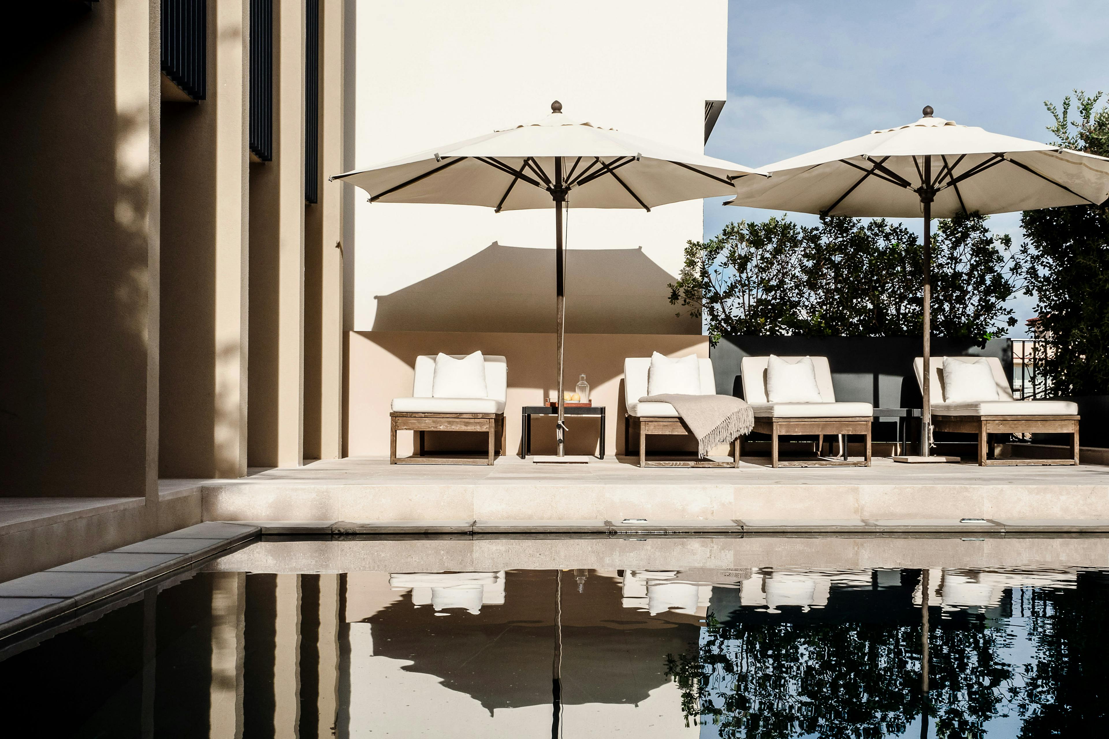 The image features a large swimming pool with a reflective surface, surrounded by a large number of white umbrellas. There are at least 12 umbrellas of various sizes and shapes, providing shade for the poolside area. The pool is also adorned with chairs and a dining table, creating a relaxing and inviting atmosphere for guests.